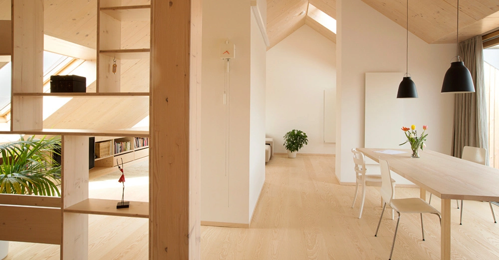 Helles Wohnzimmer mit Holzmöbeln. Stauraumregal links. Tisch mit Stühlen und Blumenstrauß rechts. Pflanze im Hintergrund. Große Fenster durchfluten den Raum mit Sonnenlicht. Holzdach verleiht rustikalen Charme.