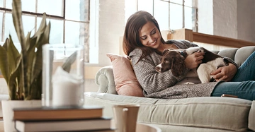 Eine Frau hält einen Welpen in ihren Armen und sitzt auf einem beigen Sofa in einem gemütlichen Wohnzimmer mit großen Fenstern im Hintergrund. Auf dem Tisch stehen Pflanzen und Bücher.