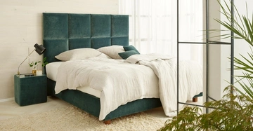 Bett mit grünem Kopfteil steht in einem modern eingerichteten Schlafzimmer mit hellen Wänden und Vorhängen. Nachttisch mit Tischlampe und Pflanze daneben. Teppich auf dem Boden.
