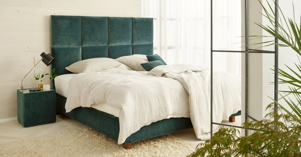 Bett mit grünem Kopfteil steht in einem modern eingerichteten Schlafzimmer mit hellen Wänden und Vorhängen. Nachttisch mit Tischlampe und Pflanze daneben. Teppich auf dem Boden.