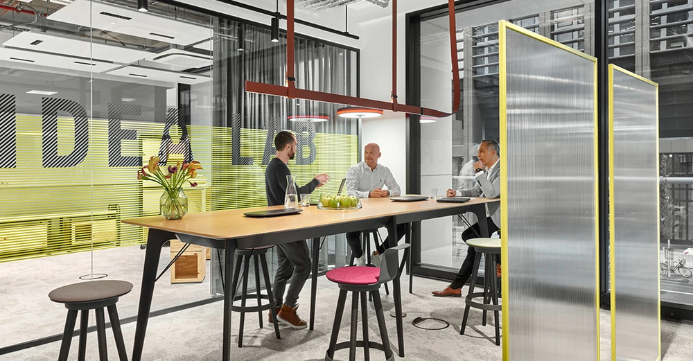 Drei Männer sitzen und unterhalten sich an einem hohen Tisch in einem modernen Büro mit großen Fenstern. Im Hintergrund steht auf einer Glaswand der Text IDEA LAB.