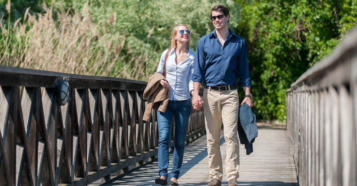 Zwei Personen gehen Hand in Hand über eine hölzerne Brücke in einem grünen, bewaldeten Parkbereich bei sonnigem Wetter.
