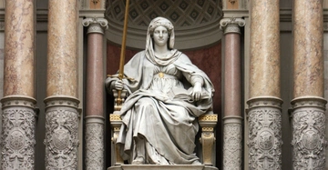 Eine Marmorstatue einer sitzenden Frau hält ein Schwert und ein Buch in einer Hand. Sie befindet sich zwischen reich verzierten Säulen in einem kunstvollen Innenraum.