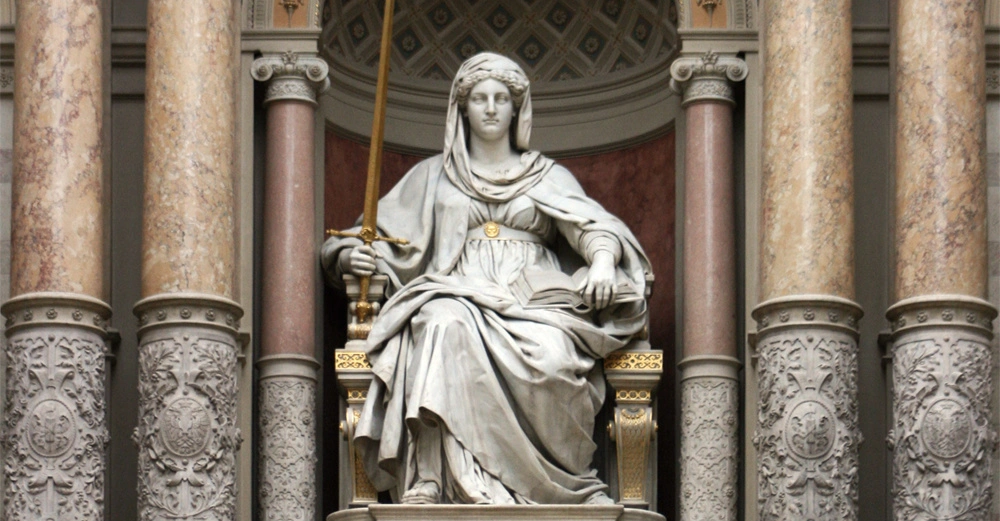 Eine Marmorstatue einer sitzenden Frau hält ein Schwert und ein Buch in einer Hand. Sie befindet sich zwischen reich verzierten Säulen in einem kunstvollen Innenraum.