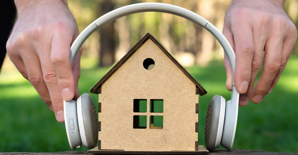 Ein hölzernes Spielzeughaus vor einem Gartenhintergrund, über das ein Kopfhörer gehalten wird.