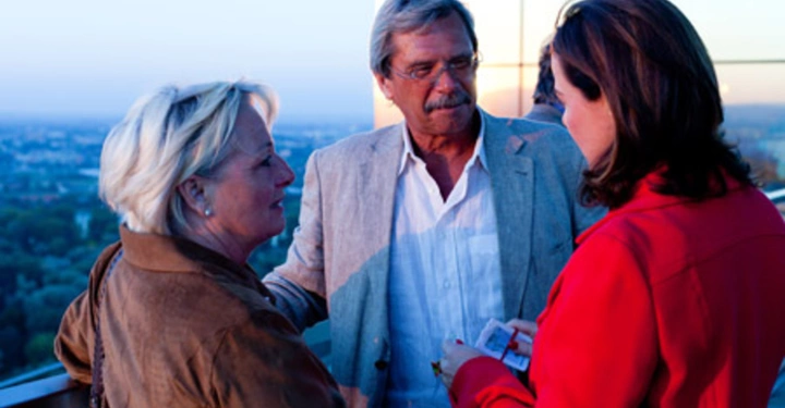 Galerie-Bild 26: Drei Personen stehen auf einem Balkon und unterhalten sich Eine Frau in einer roten Jacke hält etwas in ihren Händen Im Hintergrund sind Bäume und ein klarer Himmel zu sehen