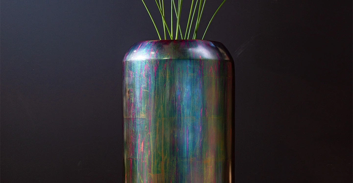 Große bunte Vase mit langen grünen Stielen und trockenen Blättern steht vor einem dunklen Hintergrund auf einem schwarzen Sockel