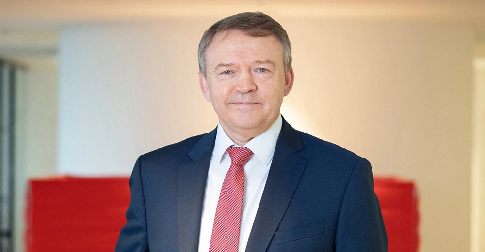 Ein Mann trägt einen blauen Anzug und eine rote Krawatte steht und lächelt in einem gut beleuchteten Raum mit roten und cremefarbenen Wänden.