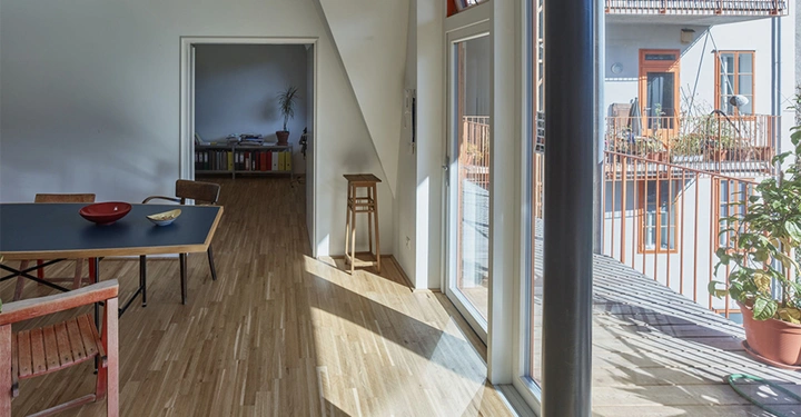 Ein Esstisch steht im Vordergrund mit einem roten und einem gelben Teller darauf. Sonnenlicht scheint durch große Fenster, die eine Terrasse und ein benachbartes Gebäude zeigen.