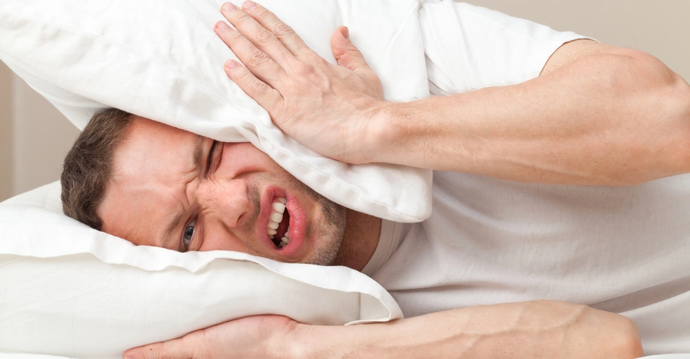 Mann liegt im Bett drückt ein Kopfkissen gegen seinen Kopf Gesichtsausdruck zeigt Unbehagen Umgebung ist ein schlichtes Schlafzimmer.