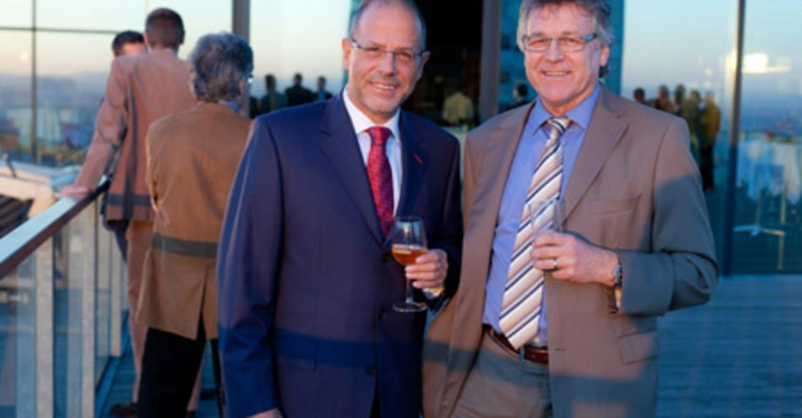 Galerie-Bild 18: Zwei Männer halten Getränke in den Händen lächelnd auf einer Terrasse eines modernen Gebäudes während des Sonnenuntergangs im Hintergrund zahlreiche Menschen in Gespräch vertieft.