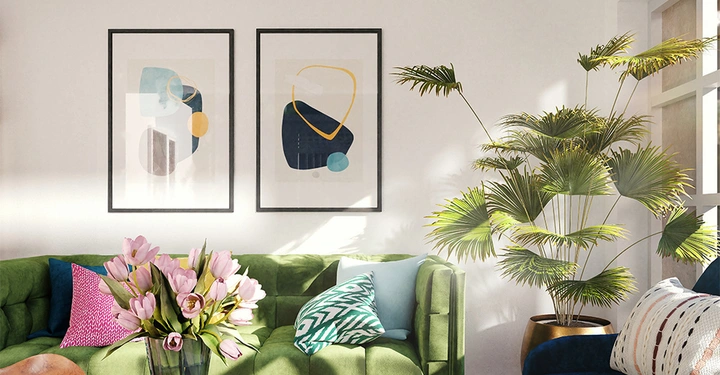 Grünes Sofa steht vor einer weißen Wand hängt mit zwei abstrakten Gemälden; eine Vase mit rosa Blumen und Kissen ist darauf platziert dann rechts daneben steht eine große Zimmerpflanze.