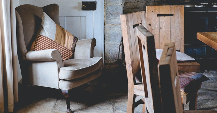 Ein gepolsterter Sessel mit Kissen steht neben einem Vorhang am Fenster in einem gemütlichen Raum mit Stühlen aus Holz und einem Tisch vor einer Steinmauer.