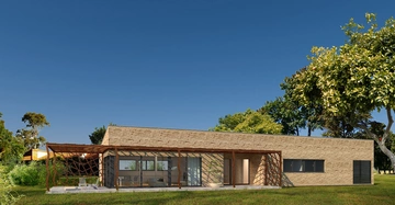 Bungalow mit Steinmauern und Holzpergola steht in einer grünen Wiese mit Bäumen im Hintergrund und klarem blauen Himmel