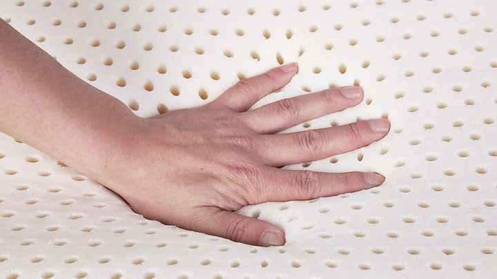 Hand drückt auf beigefarbene Latexmatratze, Lüftungslöcher sind gut zu sehen, Material wirkt fest und weich zugleich