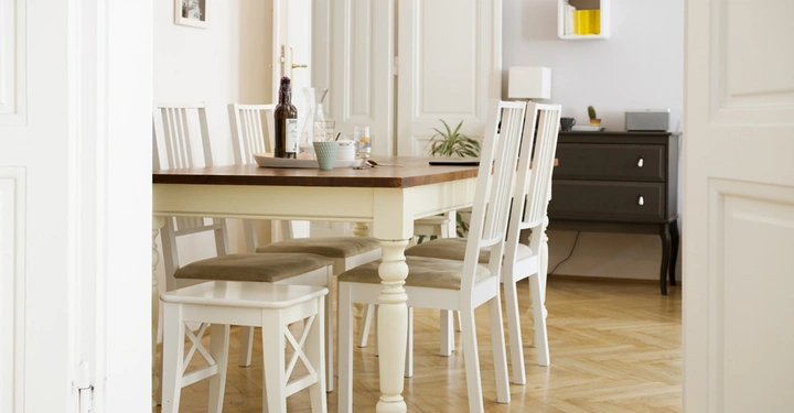 Galerie-Bild 2: Weißer Esstisch mit vier weißen Stühlen steht im Wohnzimmer auf Holzfußboden. Ein Dekotablett mit einer Weinflasche, Gläsern und einer Karaffe steht auf dem Tisch. Im Hintergrund befindet sich eine schwarze Kommode.