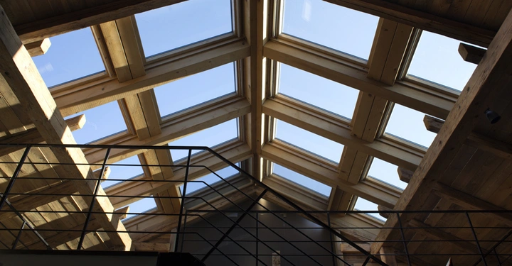 Galerie-Bild 5: Große Dachfenster lassen Sonnenlicht in einen Raum mit Holzwänden und -decke. Ein schwarzes Treppengeländer verläuft diagonal unter den Fenstern, erzeugt ein geometrisches Muster.