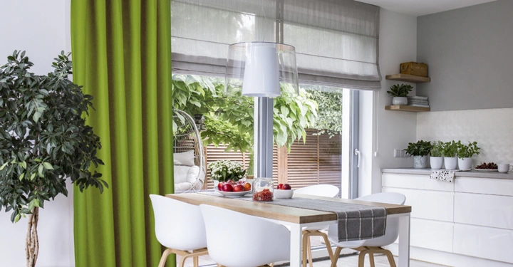 Topfpflanze steht neben grünem Vorhang im modernen Esszimmer Esszimmertisch mit vier weißen Stühlen daneben Fenster und Glastüren zur Terrasse Küchenschränke und Regale mit Pflanzen im Hintergrund