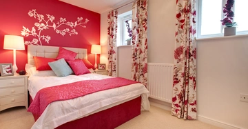 Ein Bett mit pink-weißer Bettwäsche und vielen Kissen steht in einem Schlafzimmer vor einer pinken Wand mit blumigem Wandtattoo umgeben von Vorhängen mit Blumenmuster und Nachttischen mit Lampen.