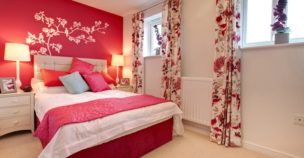 Ein Bett mit pink-weißer Bettwäsche und vielen Kissen steht in einem Schlafzimmer vor einer pinken Wand mit blumigem Wandtattoo umgeben von Vorhängen mit Blumenmuster und Nachttischen mit Lampen.