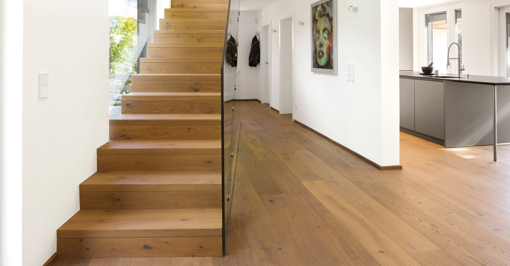 Holztreppe steigt seitlich in einem modernen, gut beleuchteten Wohnbereich empor. Daneben befindet sich eine offene Küche mit grauer Theke. Kunstwerk hängt an der weißen Wand.