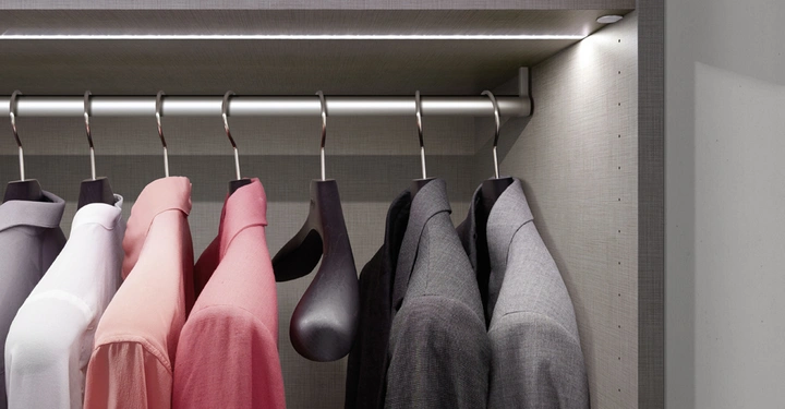Mehrere Hemden hängen ordentlich auf Kleiderbügeln an einer Kleiderstange in einem gut beleuchteten Schrank mit grauer Textur. Oben liegt gefaltete Bekleidung auf einem Regal.