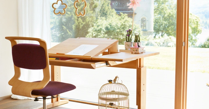 Schreibtisch mit Zeichnung sitzt vor großen Fenstern Blick auf Schlossgründe gelber Vogelkäfig unten links bunte Spielzeuge auf dem Boden Hängebloemen auf Fenster Multiple dekorative Gegenstände auf dem Tisch