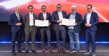 6 Männer im Anzug stehen lächelnd auf Bühne mit blau-rotem insiven Hintergrund und halten ihre Auszeichnung in die Höhe.