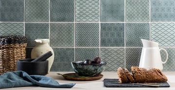 Korbflechtkorb und Mörser auf Arbeitsplatte neben einem Teller mit Pflaumen, einem Krug und Brot in einer Küche mit dekorativen Fliesen im Hintergrund.