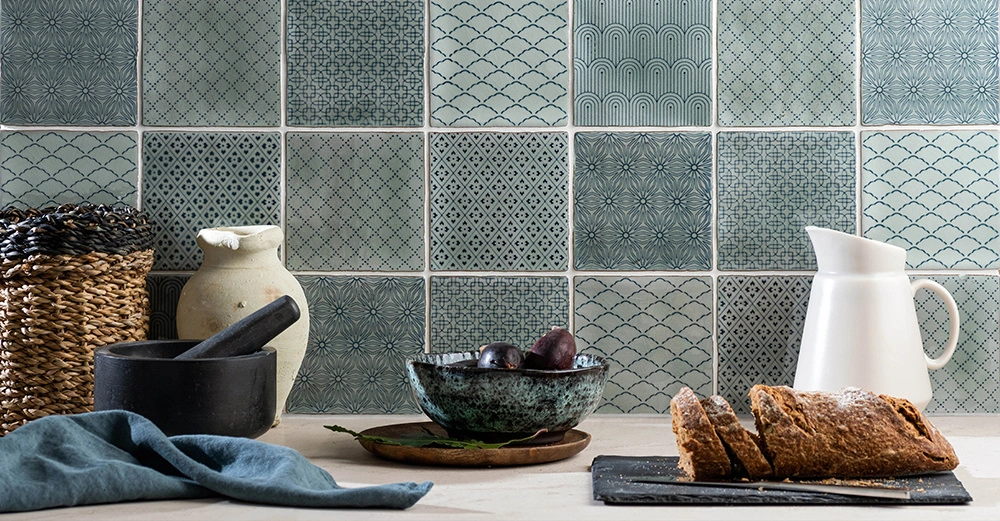 Korbflechtkorb und Mörser auf Arbeitsplatte neben einem Teller mit Pflaumen, einem Krug und Brot in einer Küche mit dekorativen Fliesen im Hintergrund.