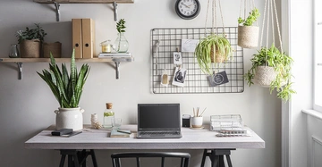 Laptop auf Schreibtisch stehend in dekorativem Home-Office-bereich mit Topfpflanzen nebenan Regale mit Büchern Pflanzen und Ordnern oben Wanduhr und Pinnwand mit Bildern und Notizen dahinter Fensternische rechts.