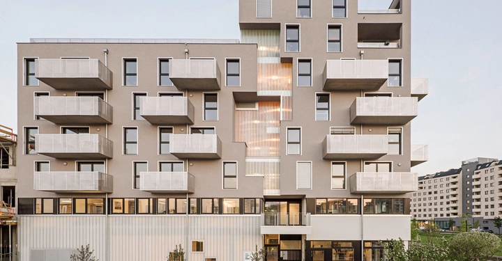 Galerie-Bild 12: Ein modernes Wohngebäude zeigt symmetrisch angeordnete Balkone und Fenster in einer städtischen Umgebung, umgeben von weiteren Gebäuden und einem Gehweg mit Bäumen im Vordergrund.