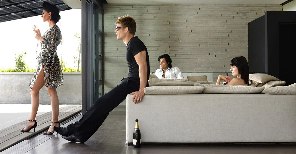 Vier Personen entspannen in einem modernen Wohnzimmer Eine Frau steht und raucht Eine andere Frau sitzt auf einem Sofa Ein Mann sitzt daneben und ein weiterer Mann sitzt hinten und lehnt sich zurück