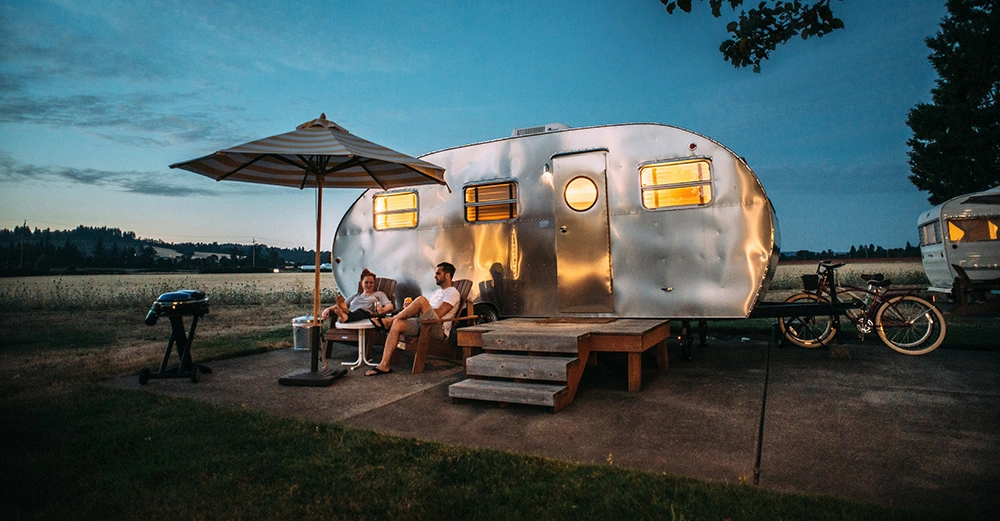 junges Paar sitzt vor seinem Retrocampingbus im Sonnenuntergang auf einem Campingplatz. Hinter dem Camper stehen zwei Fahrräder, im Hintergrund ein weiteres Wohnmobil.
