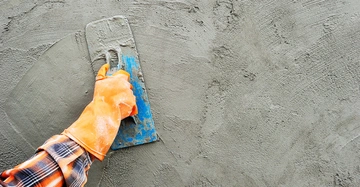 Eine Hand in einem orangefarbenen Handschuh hält eine Kelle und trägt Putz auf eine Wand auf. Im Hintergrund ist der rauhe graue Putz zu sehen.