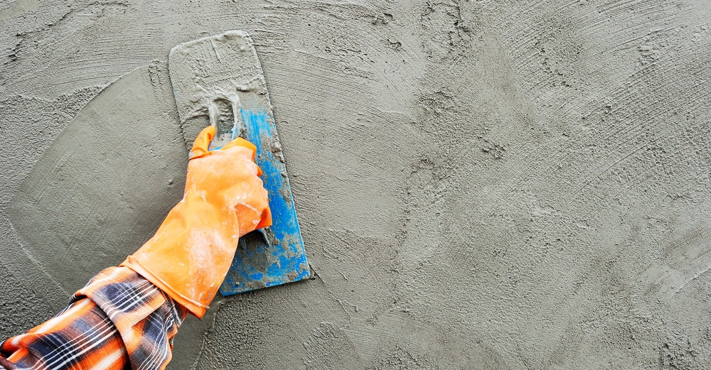 Eine Hand in einem orangefarbenen Handschuh hält eine Kelle und trägt Putz auf eine Wand auf. Im Hintergrund ist der rauhe graue Putz zu sehen.