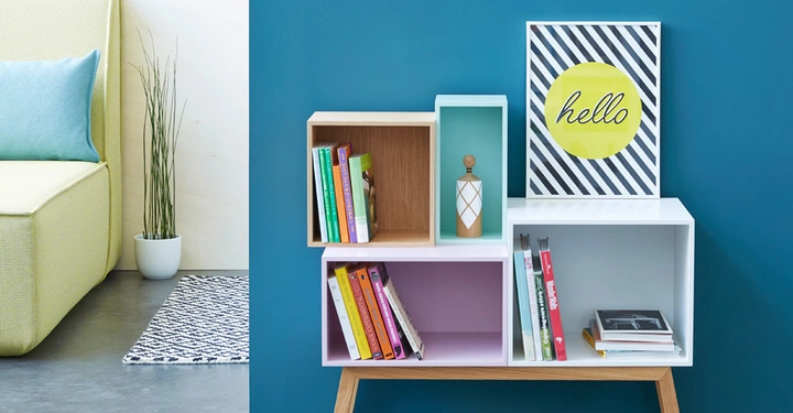 Regal mit Büchern und Dekoration steht an der blauen Wand im modernen Wohnzimmer neben einem grünen Sofa mit Kissen und einer Pflanze. Im Regal steht ein Bild mit dem Text hello.