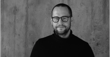 Portrait eines Mannes mit kurzem braunem Haar und markanter Brille in schwarzem Rollkragenpullover