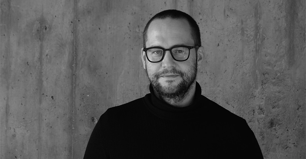 Portrait eines Mannes mit kurzem braunem Haar und markanter Brille in schwarzem Rollkragenpullover