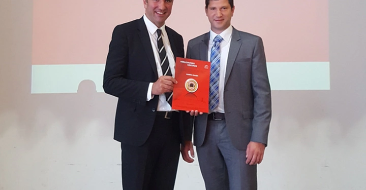 Zwei Männer stehen in Anzügen in einem Raum eine Person hält eine rote Mappe. Im Hintergrund ist eine Leinwand mit Text: 37. ordentliche Generalversammlung Österreichischer Fertighausverband