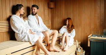 Drei Personen in weißen Bademänteln sitzen und unterhalten sich in einer holzverkleideten Sauna. Neben ihnen ein Eimer mit Schöpfkelle.