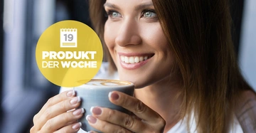 Eine Frau lächelt und hält eine Tasse mit Latte Art Kaffeetasse in beiden Händen in einem gut beleuchteten Innenraum Text auf einem gelben Kreis 19 Produkt der Woche