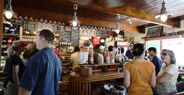 Menschen stehen an der Theke eines belebten Cafés an unterhalten sich und bestellen Essen neben Regalen und einer Tafel mit Menüangeboten die an der Wand hängt.