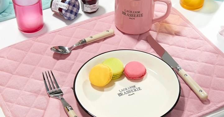 Teller mit drei farbigen Macarons liegt auf rosa Platzdeckchen umgeben von rosa Tasse Gabel Messer Löffel und kleinen Marmeladengläsern Schriftzug Lagrande Brasserie auf Tasse und Teller