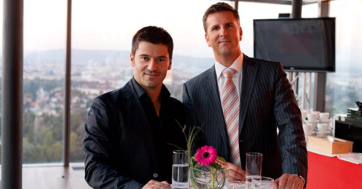 Galerie-Bild 42: Zwei Männer stehen an einem runden Tisch mit einem Blumenarrangement im Glas und Getränken Auf einer Terrasse oder in einem hoch gelegenen Raum mit Glaswänden und Blick auf eine Stadt im Hintergrund