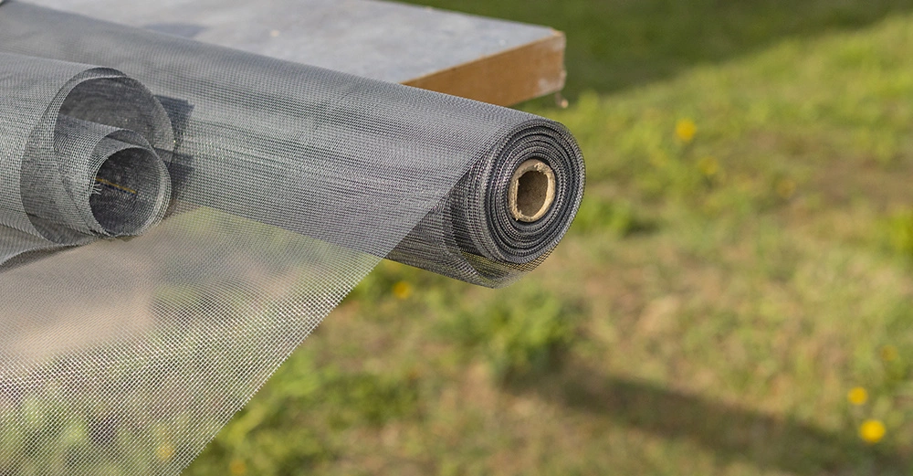 Eine Rolle schwarzes Fliegengitter liegt auf einem Holztisch im Freien, das Netz entfaltet sich und der Hintergrund zeigt eine grüne Wiese mit einigen gelben Blumen.