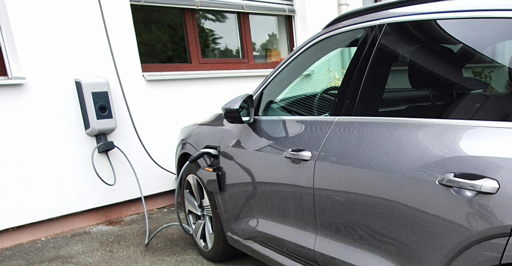 Ein graues Auto lädt an einer Wandladestation vor einem weißen Gebäude mit Fenstern mit roten Rahmen und Jalousien. Ein Ladekabel verbindet die Station mit dem Auto.