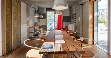 Langer Holztisch mit Stühlen und Büchern steht in modernem Raum mit großen Pendelleuchten Blick nach draußen durch Fenster auf geparkte Autos roter Vorhang im Hintergrund.