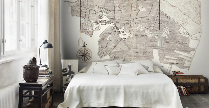 Galerie-Bild 6: Bett ruht in einem Schlafzimmer mit einem großen Kartenmuster an der Wand daneben ein kleiner Nachttisch mit Büchern Lampe und Fenster das Tageslicht hereinlässt.