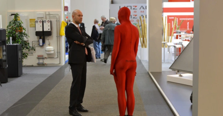 Galerie-Bild 27: Mann im Anzug spricht mit einer Person im roten Ganzkörperanzug auf einem Messegang umgeben von Ständen und anderen Besuchern